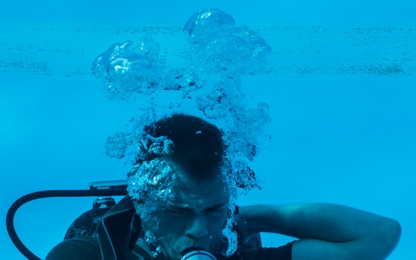 An underwater photograph of a scuba diver submerged in what appears to be a swimming pool, identifiable by the blue tiled walls visible in the background. The diver is wearing a black t-shirt, BCD (buoyancy control device), and is breathing through a regulator, producing a large cluster of air bubbles rising above their head. The image captures a likely training or certification exercise scenario.
