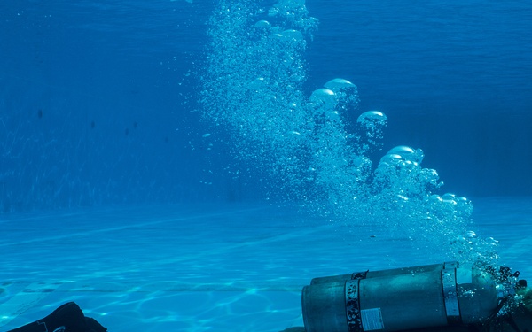 An underwater photograph captures a scuba diver near the bottom of a swimming pool. The diver is in a crouched or kneeling position on the pool floor, wearing full scuba gear including a tank, BCD, mask, and fins. A trail of exhaled air bubbles rises prominently toward the water surface above.