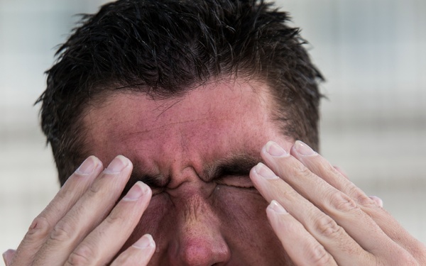 A close-up photograph of an adult male pressing both hands against his face, covering his eyes. His facial expression and flushed, reddened skin suggest distress, pain, or intense emotional strain. The background is blurred, keeping full focus on the subject.