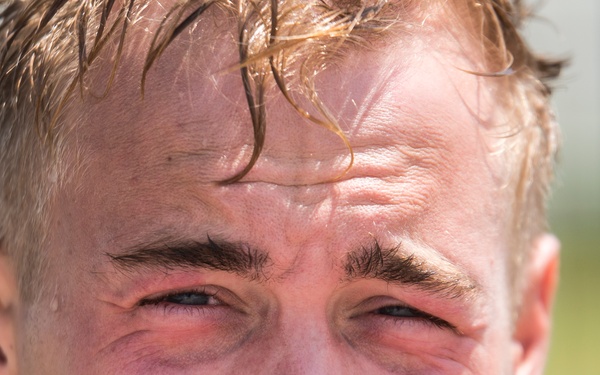 Close-up portrait of a young adult male with short blonde hair that appears wet or sweaty. His eyes are visibly red and irritated, and his skin is flushed, suggesting recent physical exertion or exposure to an irritant. He is wearing what appears to be a dark military or tactical-style garment.