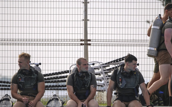 A group of individuals in scuba diving gear are gathered at the edge of an outdoor swimming pool. Four people are seated at the pool's edge wearing buoyancy control devices (BCDs) and tanks, while two others are standing and walking nearby. The setting appears to be a structured diving training session at a facility with chain-link fencing.