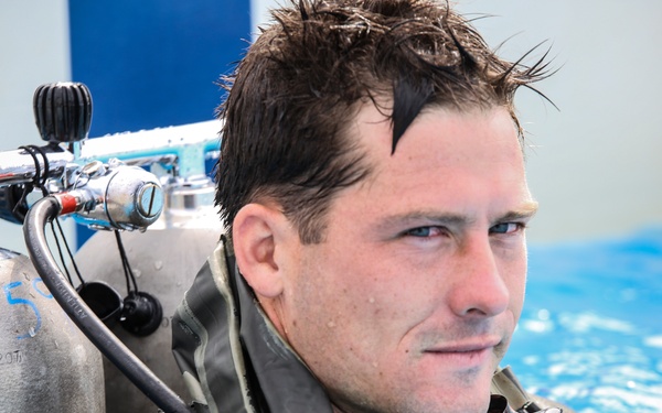 A man wearing full scuba diving gear is partially submerged in what appears to be an indoor swimming pool. He is equipped with a tank, regulator hoses, and a wetsuit, with wet hair suggesting recent submersion. He appears calm and is looking slightly off-camera.