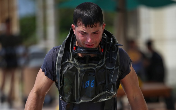 A young man is walking while soaking wet, wearing a blue t-shirt and carrying diving or underwater breathing apparatus equipment around his neck marked with '106'. He appears physically exhausted, with his head down and clothes completely drenched, suggesting he has just completed a water-based training exercise or activity. The background shows an outdoor facility with other individuals and structures visible but out of focus.