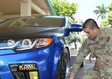 A U.S. Army soldier in MultiCam (OCP) camouflage uniform is crouched down detailing or cleaning a heavily modified blue Kia Forte Koup. The vehicle features aftermarket modifications including a carbon fiber hood, lowered suspension, custom wheels, yellow fog lights, and a 'KDM Registry' front license plate. The setting appears to be a covered parking area on what looks like a military installation, with palm trees visible in the background.