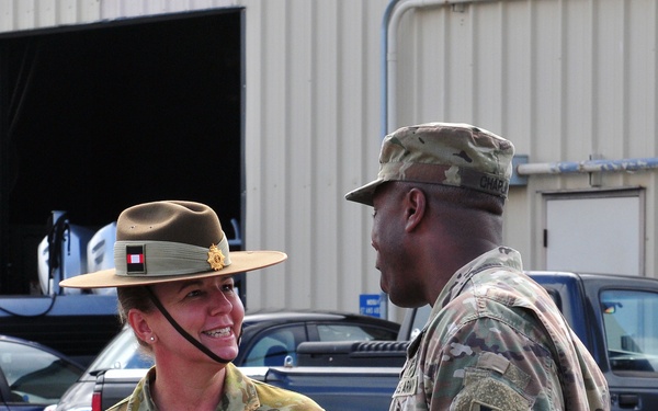 Two military personnel in camouflage uniforms are engaged in conversation outdoors near a building and parked vehicles. The individual on the left is wearing an Australian Army uniform with a distinctive wide-brimmed slouch hat and a name tape reading 'FELMINGHAM', while the individual on the right is wearing a US Army uniform with an OCP pattern and a unit patch visible on the left sleeve. The interaction appears cordial and informal, suggesting a liaison or joint military engagement.