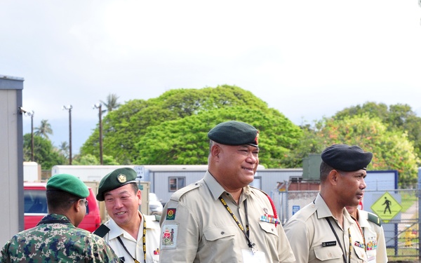 The image shows a group of military officers in uniform gathered in an outdoor paved area, likely at a military installation or event. Several officers wearing tan/khaki dress uniforms with green berets and medal ribbons are present, along with one soldier in digital camouflage. A handshake greeting is taking place between two individuals, suggesting a formal reception or multinational military gathering.