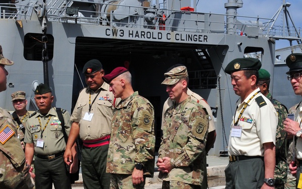 A group of military personnel from multiple nations are gathered on a dock in front of a U.S. Army vessel named 'CW3 Harold C Clinger.' The group appears to be engaged in a briefing or orientation, with one U.S. soldier in multicam uniform facing the others. The diverse uniforms suggest this is a multinational military exercise or official visit.