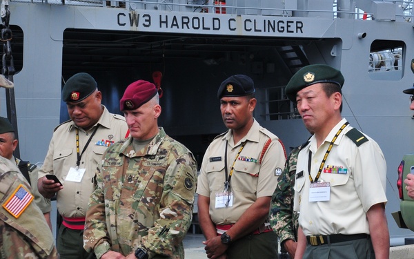 A group of military personnel from multiple nations are gathered on a dock in front of the US Army vessel 'CW3 Harold C Clinger.' A US Army soldier in MultiCam uniform appears to be briefing or greeting the international delegation. The group includes officers wearing various national uniforms, berets, and service ribbons.