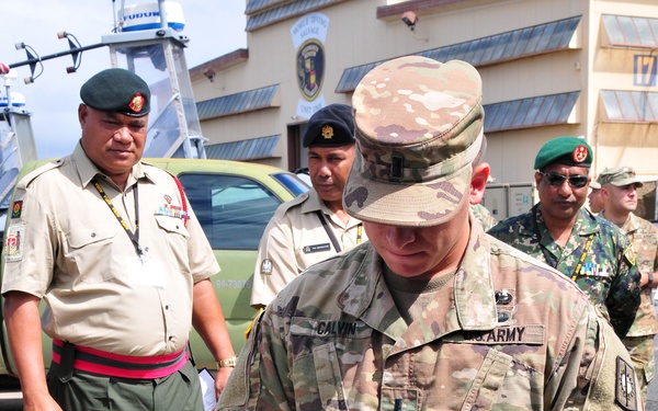A U.S. Army soldier named Calvin is operating a game-style controller connected to a ruggedized military laptop labeled 'MTRS MK1 MOD1', likely demonstrating a remote-controlled system. Several multinational military officers in various uniforms are observing the demonstration in an outdoor setting near a military facility. The gathering appears to be a multinational military exercise or equipment demonstration.