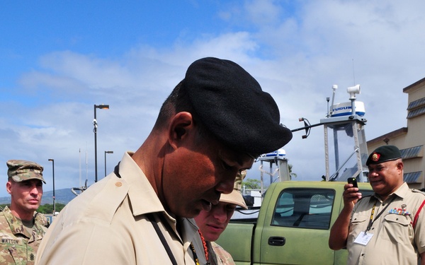 A group of military personnel from multiple nations are gathered outdoors, likely at a military installation or port facility. The central figure, wearing a tan uniform with a black beret and a shoulder patch depicting a coat of arms, is examining a handheld blue electronic device connected by a wire. The scene appears to be a demonstration or equipment familiarization event, with observers in various military uniforms watching.