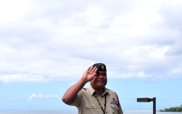 A senior military officer in a tan/khaki uniform with a black beret, medal ribbons, and a distinctive red-and-black striped belt is rendering a salute while standing on what appears to be a military dock or pier structure near the ocean. He is wearing a visitor/conference lanyard with a name badge. Two other personnel in US Army camouflage uniforms are visible in the background.