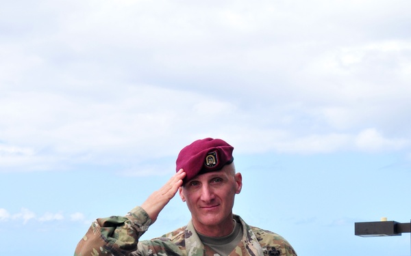 A U.S. Army soldier wearing a maroon beret and OCP (Operational Camouflage Pattern) uniform is rendering a salute while standing on what appears to be a military watercraft or dock structure near open water. His name tape reads 'FERRUSI' and he wears rank insignia consistent with a Sergeant First Class or higher NCO grade. A second soldier in the background appears to be working on equipment.