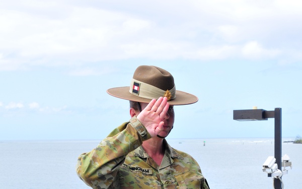 An Australian Army officer in Multicam (AMCU) camouflage uniform is standing on a pier or dock near open water, rendering a salute while holding what appears to be a presentation case or award box. A second military officer in a different uniform (olive green, likely from another nation) is visible in the lower left. The setting appears to be an outdoor military ceremony at a waterfront location.