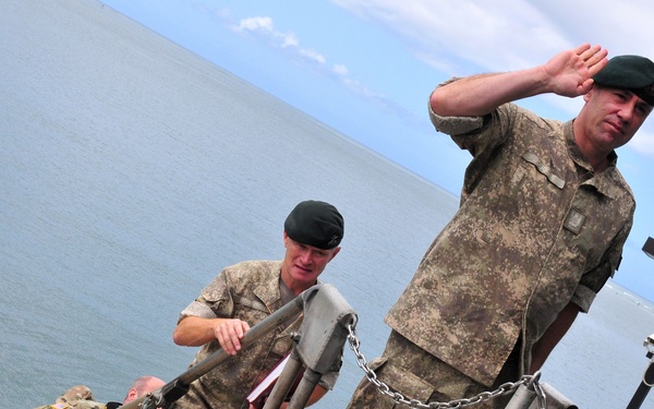 Military personnel in camouflage uniforms are ascending or standing on what appears to be a military vessel or floating platform near a coastline. The foreground soldier is rendering a salute while wearing a dark green beret, suggesting a formal military greeting or acknowledgment. Multiple soldiers from what appear to be different national militaries are present, indicated by varying uniform patterns.