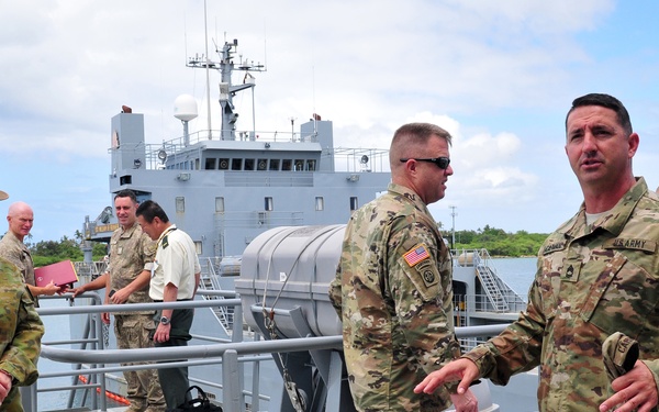 A multinational military gathering is taking place on the deck of a gray naval vessel, likely during a joint exercise or official visit. Personnel from at least two identifiable nations are present, including US Army soldiers in MultiCam/OCP uniforms and an Australian Army officer in DPCU uniform with a distinctive wide-brimmed slouch hat. The setting appears to be a tropical or subtropical port environment based on the vegetation and weather conditions.