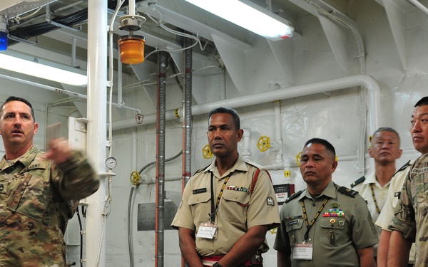 A U.S. Army soldier appears to be conducting a briefing or tour for a multinational military delegation inside what appears to be a ship's engine room or mechanical space. The group includes personnel in U.S. Army combat uniforms and foreign military officers wearing tan and olive green uniforms with name badges/lanyards. The setting suggests an official military exchange, inspection, or cooperative engagement event.