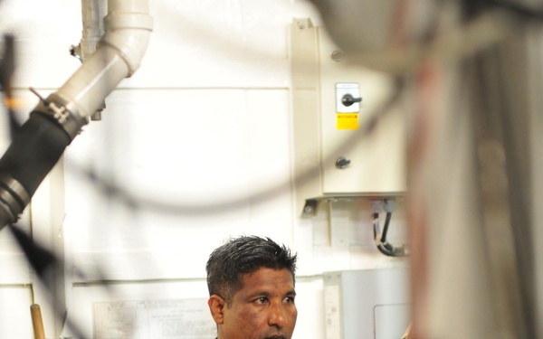 A military officer named 'AHMED' wearing digital camouflage uniform with a Maldives flag patch on the sleeve stands in what appears to be an industrial or military facility. He is wearing service ribbons/medals on his chest and an event lanyard/badge, suggesting attendance at a military conference or exhibition. Other uniformed personnel are partially visible in the foreground and background.