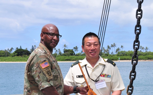 Two military personnel are shaking hands on what appears to be a naval vessel or dock, with a tropical waterway and palm tree-lined shore visible in the background. One individual wears U.S. Army multicam combat uniform with an American flag patch and airborne tab, while the other wears a light-colored military dress uniform with ribbons and a visitor badge. The person in the dress uniform is holding a wooden award or commemorative plaque shaped like a paddle or ukulele.