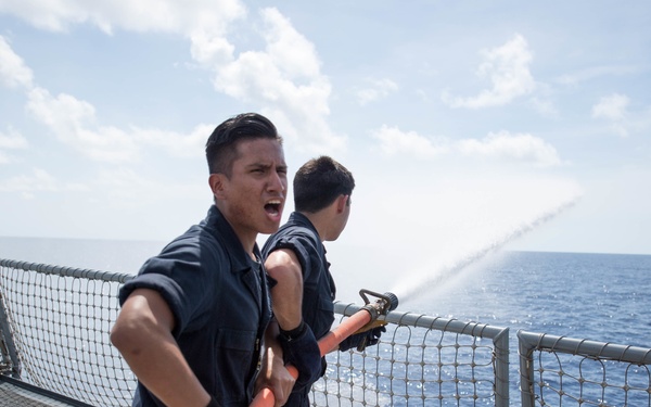 Two naval personnel in dark blue coveralls are conducting a firefighting drill or washdown exercise on the deck of a military vessel at sea. The foreground sailor is holding an orange fire hose and appears to be shouting commands or communicating, while a second sailor behind him directs the water stream over the ship's railing into the ocean. The scene takes place on a clear, sunny day with calm blue waters visible in the background.