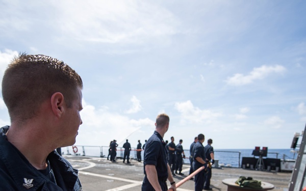 A group of naval personnel in dark blue uniforms are working on the deck of a military vessel at sea. The foreground sailor is holding an orange hose or line, while other crew members are visible in the background engaged in deck operations. The scene appears to depict a routine seamanship or replenishment evolution aboard a U.S. Navy warship.