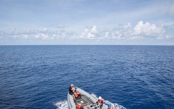 A rigid-hull inflatable boat (RHIB) carrying approximately 8-10 personnel in red life vests and colored safety helmets is underway on open ocean, departing from or operating alongside a larger naval vessel visible on the left edge. The personnel appear to be military or coast guard crew conducting a boat operations drill or transit. The sea conditions are calm with deep blue water and partly cloudy skies.