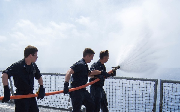 Four naval personnel in dark blue uniforms are conducting a firefighting drill on the deck of a military vessel at sea. They are operating an orange fire hose, directing a powerful stream of water over the ship's railing toward the ocean. The exercise appears to be a standard damage control training evolution performed on an open ocean deployment.