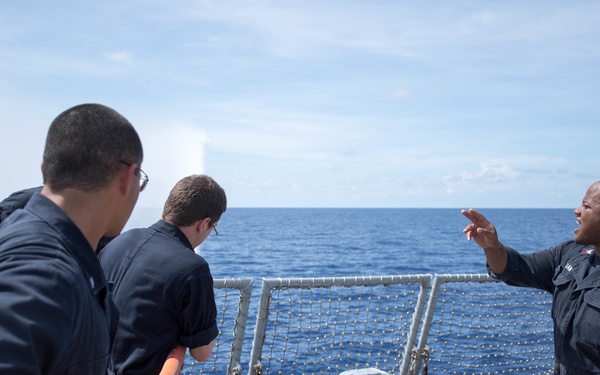 Several U.S. Navy sailors are on the deck of a naval vessel at sea, engaged in what appears to be a training or operational drill. One sailor on the right is gesturing and giving directions to the others, one of whom is holding an orange-handled tool or equipment. The scene takes place on an open ocean under partly cloudy skies.