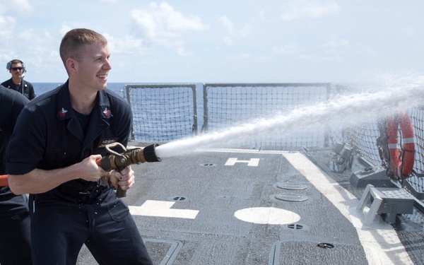 Two naval personnel in dark blue uniforms are conducting a firefighting drill on the deck of a military vessel, operating a high-pressure fire hose together. A third crew member is visible in the background wearing headphones. The scene takes place on what appears to be a helicopter deck (marked with 'H' and circle) of a naval warship at sea.