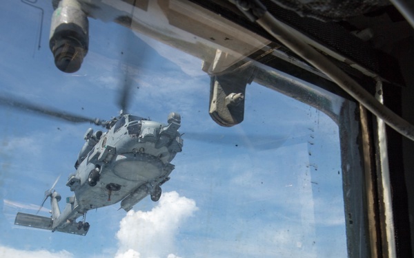 A military helicopter, appearing to be a Sikorsky MH-60 Seahawk, is photographed through a dirty window from what appears to be a ship's interior or control station. The helicopter is hovering or in low flight over what looks like a naval vessel's deck. The photo is taken from an interior vantage point looking outward through glass.