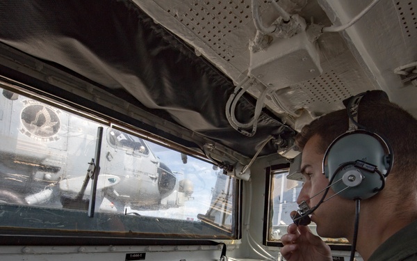 A military personnel member wearing a headset and olive drab flight suit is seated at a control station, appearing to communicate via a boom microphone. The operator is looking through windows at military aircraft on what appears to be a flight deck or airfield. The control panel in front of him contains labeled systems including 'DECK STATUS,' 'BRIDGE INFORMATION,' and 'RSD BEAM POSITION,' suggesting a naval aircraft carrier or similar military vessel.