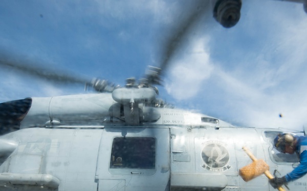 A naval deck crew member is placing or removing wheel chocks from a military helicopter on what appears to be a ship's flight deck. The helicopter, numbered '704', is a gray naval rotary-wing aircraft with rotor blades in motion. The low-angle, wide-lens perspective creates a dramatic composition emphasizing the scale of the aircraft and the urgency of the deck operation.