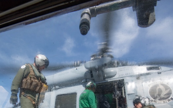 The image is taken through a glass window or canopy, showing military personnel on what appears to be a naval vessel flight deck alongside a gray military helicopter with tail number '704'. Three individuals are visible: one in full flight gear and helmet, one in a green shirt, and one crouching in olive flight suit. The helicopter displays a squadron insignia on its fuselage and has rotor blades visible at the top of the frame.