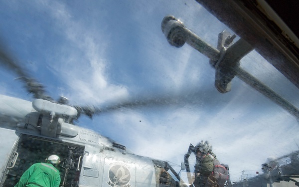 The image is taken through a glass or plexiglass window on what appears to be a naval vessel's flight deck, showing a military helicopter (likely an MH-60 Seahawk) with its rotors spinning. Personnel in flight gear and deck crew uniforms are visible around the aircraft. The photo is shot from an interior vantage point, creating reflections and distortion through the transparent surface.
