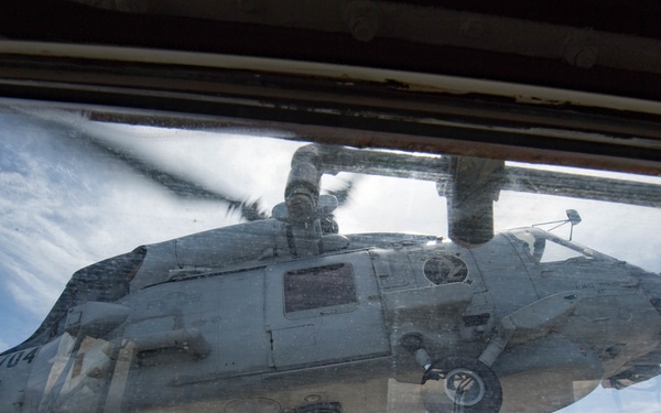 A military helicopter, marked with the number '704', is captured hovering or landing on what appears to be a naval vessel's flight deck, photographed through a window or porthole. The image is taken from a low angle through glass, giving a unique perspective of the aircraft from below. The setting appears to be at sea, with ocean and partly cloudy skies visible in the background.