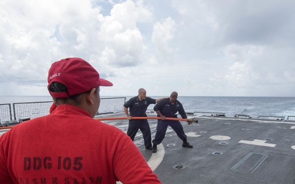 The image shows sailors aboard a U.S. Navy destroyer, identified as DDG 105 (USS Dewey), conducting deck operations on what appears to be the helicopter flight deck. Personnel are engaged in a line-handling or equipment drill, with one supervisor in a red shirt overseeing the activity. The ship is underway at sea under overcast skies.