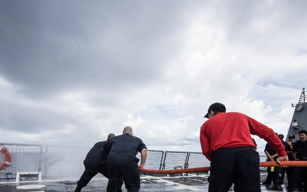 Naval personnel are conducting a firefighting drill or hose exercise on the deck of a military vessel at sea. Crew members in dark uniforms and one in a red sweatshirt are handling a large orange fire hose, with water visibly discharging at the forward end of the deck. The overcast sky and wet deck surface suggest active maritime conditions during the exercise.