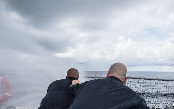 Two naval personnel in dark blue coveralls are operating a fire hose on the deck of a military vessel at sea. They are directing a high-pressure water stream over the side of the ship, likely conducting a damage control drill or washdown exercise. The scene takes place under overcast, stormy-looking skies with open ocean visible in the background.