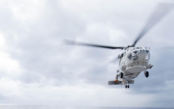 A military helicopter is captured in the process of landing or taking off from the flight deck of a naval vessel at sea. The helicopter, painted white with visible markings including the number '414', hovers just above the marked landing pad. The overcast sky and open ocean are visible in the background, suggesting open-water operations.