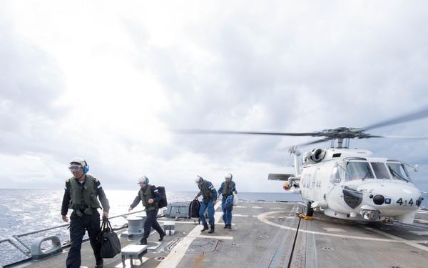 Military personnel are walking away from a white helicopter on the flight deck of a naval vessel at sea. The helicopter, marked with the number '414', appears to have just landed or is preparing for departure, with its rotors still spinning. The scene takes place under overcast skies with open ocean visible in the background.