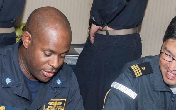 Two military officers in naval uniforms are seated at a table, signing documents together. The officer on the left wears a US Navy uniform with a name tag reading 'ANTHONY WENDY' and the title 'COMMANDING OFFICER,' while the officer on the right wears what appears to be an East Asian navy uniform with rank insignia. The scene appears to be a formal signing ceremony, likely representing a bilateral or cooperative naval engagement.