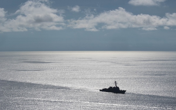 An aerial photograph taken from an aircraft showing a naval warship underway on a calm, sunlit open ocean. The ship appears to be a destroyer or frigate-class vessel, visible as a dark silhouette against the reflective silver water surface. The image is captured from a low-altitude aircraft, with part of the aircraft's wing or fuselage visible in the lower-left corner.