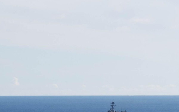 Two naval vessels are underway together on open blue ocean under a partly cloudy sky, suggesting a joint maritime exercise or transit operation. The larger vessel on the left appears to be a flat-deck helicopter destroyer or light carrier, while the smaller vessel on the right appears to be a guided-missile destroyer. Both ships are leaving visible wakes, indicating active forward movement.