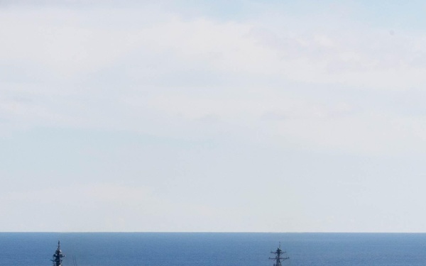 The image shows a naval formation of three warships underway on open ocean under partly cloudy skies. A large flat-deck helicopter destroyer or light carrier is prominently visible in the foreground-left, accompanied by what appears to be an Arleigh Burke-class guided-missile destroyer to its right, and a third smaller vessel visible on the distant horizon. The formation suggests a joint or allied naval exercise or transit operation.