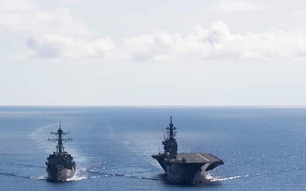 Two military naval vessels are sailing in formation across open blue ocean waters. The ships consist of what appears to be a destroyer-class warship on the left and a larger flat-deck vessel (likely a helicopter destroyer or light carrier) on the right. Both vessels are underway, leaving visible wakes in calm sea conditions under a partly cloudy sky.