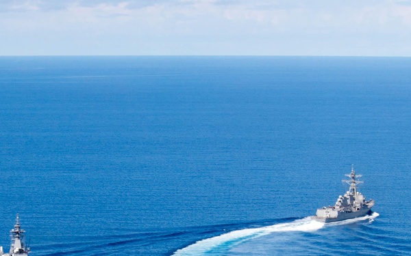 An aerial photograph shows two naval vessels underway in open blue ocean waters. The larger vessel on the left appears to be an amphibious assault ship or light aircraft carrier with a flat flight deck, while the smaller vessel on the right appears to be a guided-missile destroyer or cruiser. The destroyer is executing a turning maneuver, leaving a prominent curved white wake as it repositions relative to the larger ship.