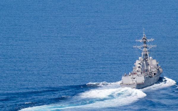 An aerial photograph captures a gray naval warship, consistent with a U.S. Navy Arleigh Burke-class guided-missile destroyer, underway at speed on open blue ocean. The vessel is executing a turn, evidenced by the curved white wake trailing behind it. The image is taken from an elevated angle, likely from an aircraft or another vessel.