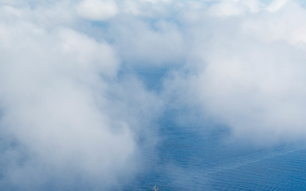 An aerial photograph captures a single naval warship traversing open ocean, leaving a visible white wake behind it. The vessel appears small relative to the vast expanse of deep blue water surrounding it. A dense bank of low-lying clouds occupies the upper portion of the frame, creating a dramatic contrast with the ocean surface.