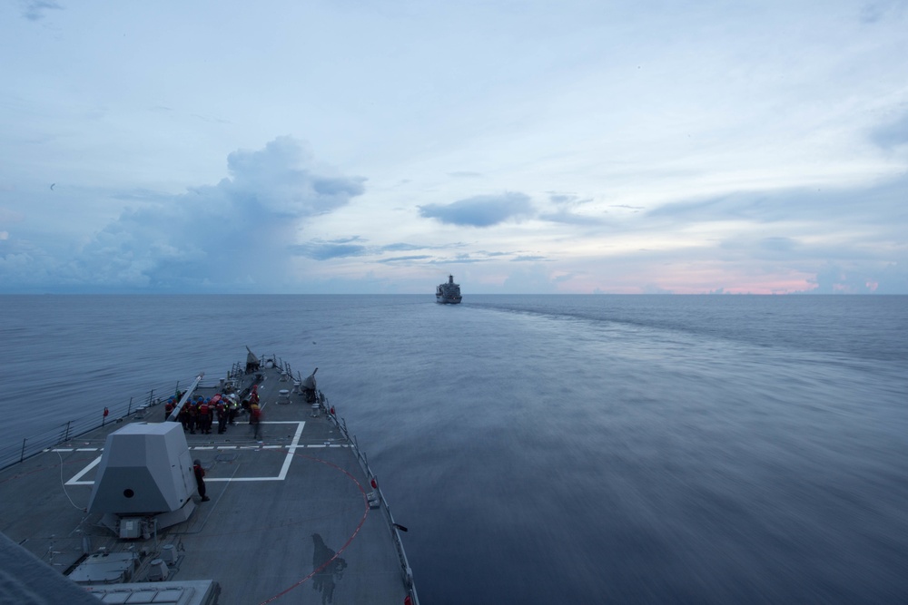 The image shows the bow of a naval warship, likely a guided-missile destroyer, photographed from an elevated forward position looking ahead across calm open ocean. A second vessel is visible in the distance on the horizon, possibly a support or replenishment ship. The scene appears to be taken at dusk or dawn, with a partially cloudy sky showing soft pink and blue tones.