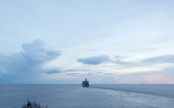The image shows the bow of a naval warship, likely a guided-missile destroyer, photographed from an elevated forward position looking ahead across calm open ocean. A second vessel is visible in the distance on the horizon, possibly a support or replenishment ship. The scene appears to be taken at dusk or dawn, with a partially cloudy sky showing soft pink and blue tones.