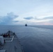 The image shows the bow of a naval warship, likely a guided-missile destroyer, photographed from an elevated forward position looking ahead across calm open ocean. A second vessel is visible in the distance on the horizon, possibly a support or replenishment ship. The scene appears to be taken at dusk or dawn, with a partially cloudy sky showing soft pink and blue tones.